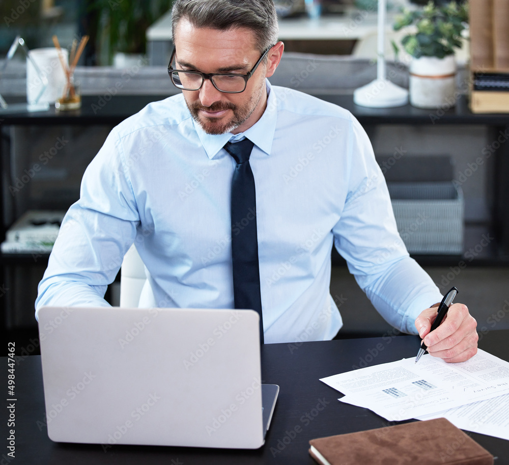 Poster his work is a priority. shot of a mature businessman using a laptop while going through paperwork in