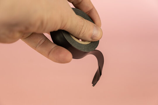 An adult male hand holding black electrical tape against a pink background. Electrical tape is used to insulate electrical wires and other materials that conduct electricity.