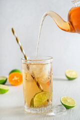 Refreshing homemade organic iced tea in drinking glass with ice cubes, sour lime fruit slices and paper straw with liquid pouring from teapot served on white wooden table at kitchen in summer