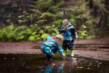 Two little boys are playing in a muddle puddle. There is a reflection in a puddle. Image with selective focus - obrazy, fototapety, plakaty