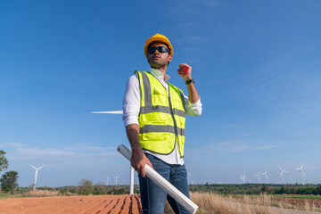 Engineer wearing uniform ,helmet hold document inspection work in wind turbine farms rotation to generate electricity energy. Green ecological power energy generation wind sustainable energy concept.