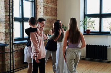 Group of happy teenage guys and girls in activewear giving hugs to one another before repetition in...