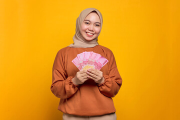 Smiling young Asian woman in orange shirt holding money banknotes isolated over yellow background