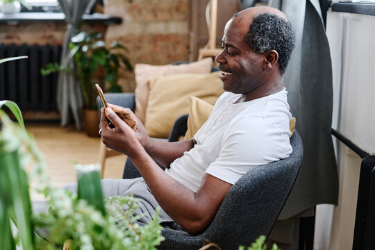 Contemporary senior black man in home wear communicating in video chat while sitting in comfortable armchair in living room