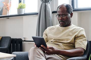 Serious retired man in eyeglasses and home wear looking at screen of tablet while watching online...