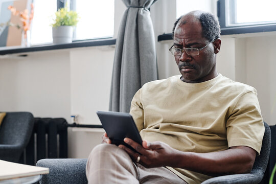 Serious retired man in eyeglasses and home wear looking at screen of tablet while watching online movie or other curious video