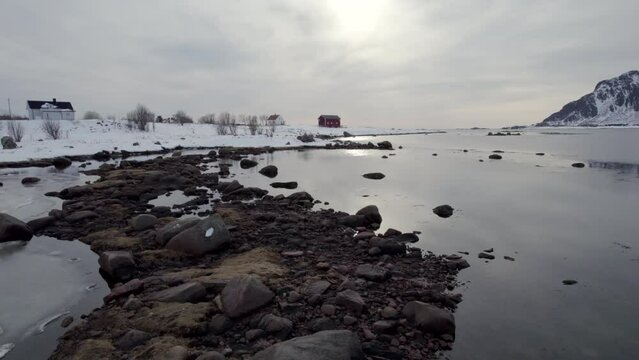 Flying over froozen ocean, rocks and water with the sun reflecting on the surface, revealing a Norwegian Farm.