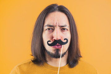 Movember concept - young frowned man holds fake paper moustache on a stick yellow background copy...