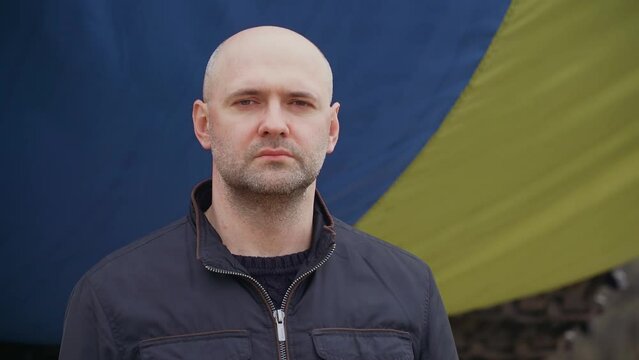 Sad refugee man standing near big waving Ukrainian flag in foreign country during invasion. Crisis, peace, stop aggression in whole world