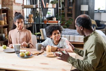 Fototapeta na wymiar Contemporary retired black man talking to his grandchildren by table served with peanut butter, bread, apples and tea for breakfast