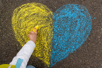 A child's hand draws a heart with crayons on the pavement with the patriotic national Ukrainian colors of yellow and blue. Stop the war in Ukraine.