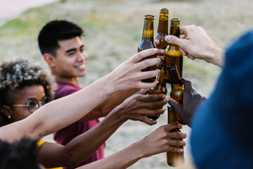 Group of multiracial friends toasting with bottles of beer - Young people celebrating together drinking with alcohol beverage on summer party - Focus on bottle and hand
