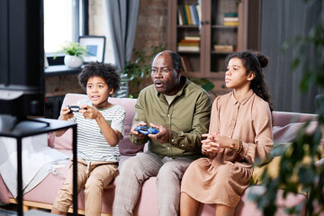 Contemporary African American grandfather and grandson with joysticks playing leisure game on couch while serious girl sitting near by