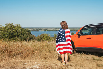 A young woman with an American flag stands near her car in nature. View from the back.