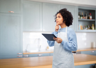 cooking, culinary and people concept - thinking woman in apron with tablet pc computer over home kitchen background