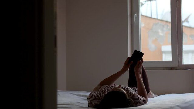 A woman lies on the bed and reads messages on the phone, the girl writes in instant messengers, the girl is in the bedroom