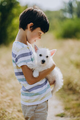 Friendship of a child with a dog. Happy boy holding a West Highland White Terrier puppy in his arms. The child walks with his puppy in the summer in the park.
