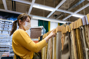 A woman with a mask on her face stands in a fabric store and selects from samples on the counter during the COVID-19 pandemic