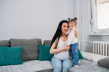 A blonde daughter with ponytails stands on the sofa next to her pregnant mother,while the child-bearing caucasian woman sits.They hug each other and look at the camera.Mother holds her daughter's hand