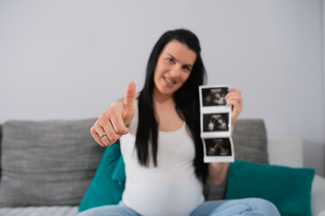 Pretty caucasian pregnant female shows ultrasound pictures of her unborn baby and at the same time with her right hand, she shows OK with raised tumb. A smiling woman looks right at the camera.