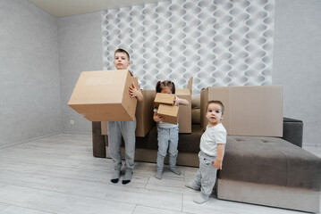 Brothers and sister are playing and enjoying moving to a new apartment against the background of cardboard boxes and a sofa. Purchase of real estate. Housewarming, delivery.