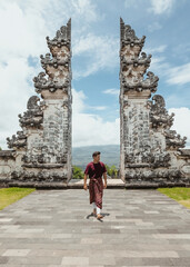 young male tourist wearing a traditional balinese sarong walking in front of gates of heaven temple...