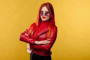 Glamour woman wearing sunglasses in red leather jacket and with red hair posing on studio background