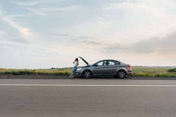 Fototapeta premium A young girl stands near a broken-down car in the middle of the highway during sunset and tries to call for help on the phone. Waiting for help. Car service. Car breakdown on road.