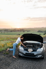 A young girl stand near a broken car in the middle of the highway during sunset and tries to repair it. Troubleshooting the problem. Car service. Car breakdown on the road.