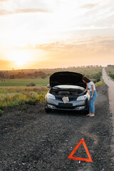 Obraz na płótnie Canvas A young girl stands near a broken car in the middle of the highway during sunset and tries to repair it. Troubleshooting the problem. Waiting for help. Car service. Car breakdown on road..