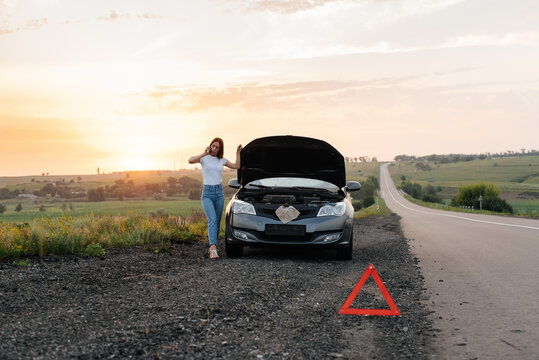 A Young Girl Stands Near A Broken Car In The Middle Of The Highway During Sunset And Tries To Call For Help On The Phone And Start The Car. Waiting For Help. Car Service. Car Breakdown On The Road.