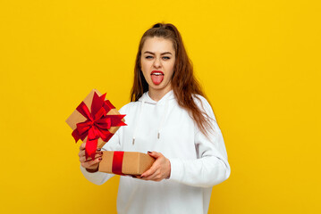 Curious brunette female opens box and looking with scorn and disappointment at gift, stands on yellow background. Holiday concept