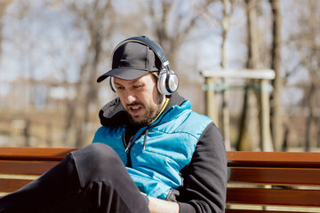 A man wearing headphones relaxes after a run, sits on a public park bench, uses phone, reads the news, turns on favorite music.