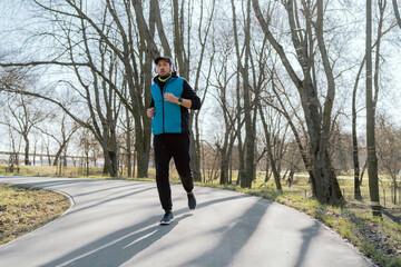 Fit, bearded young man jogging in the park, listening to music through wireless headphones, morning workout for physical health, relaxation in the form of exercise.