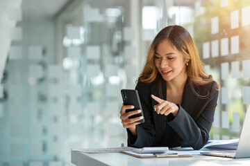 Young beautiful asian woman using smartphone and working with laptop while sitting at office desk, working from home concept.