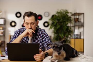A thoughtful brunet looks at a latpotp, reading papers sent by his employer. The man works remotely from his office. A modern living room can be seen in the background.