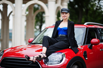 Fototapeta na wymiar Portrait of beautiful blonde sexy fashion woman model in cap and in all black with bright makeup sit on hood of red city car.