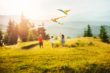 Three children running towards sun in mountains at sunset. Kids play with kites. Happy summer vacation and healthy lifestyle concept. Sunlit mountain ranges with coniferous trees. - Powered by Adobe