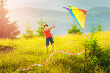 Little school boy with two hands holds a huge multicolored kite with a long, winding tail on a lace. Sunny summer or spring day at sunset.