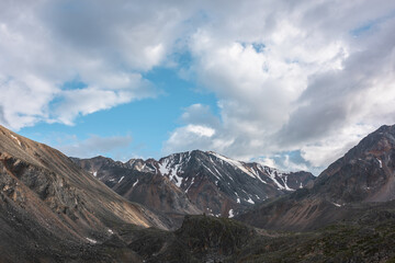 Scenic landscape with high snow mountain with sharp rocky pinnacle in sunlight under clouds in blue sky at changeable weather. Colorful mountain scenery to sunlit large mountain top under cloudy sky.