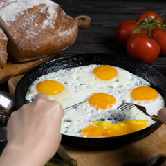 Cooked breakfast - scrambled eggs in pan, tomatoes, bread kitchen table. Female hands with knife and fork