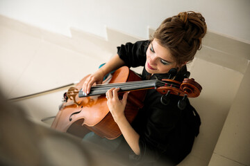 female cellist sits on stairs and plays cello with fingers, pizzicato technique, enjoys performing music, top view, suitable for poster or poster - Powered by Adobe