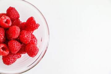 Top view of bright juicy raspberries in half of a glass bowl on left side of white table background, for health eating and lifestyle