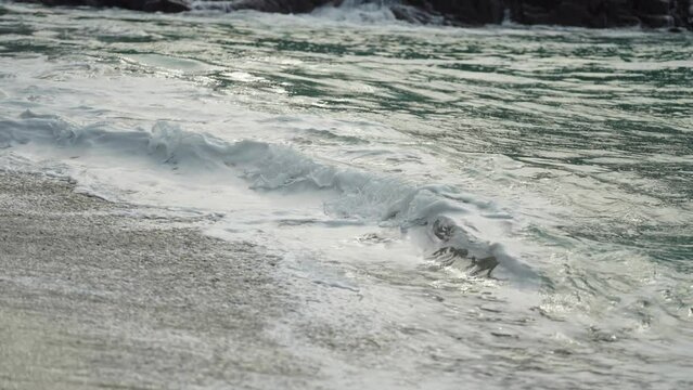 A close-up shot of the long waves spilling on the sandy beach. Slow-motion.