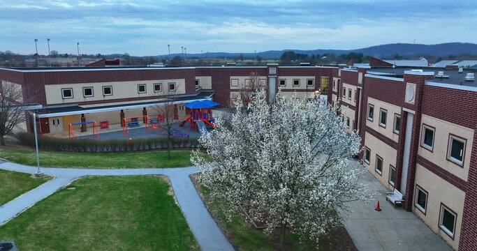 School in USA and student recess playground. Aerial past blooming tree in spring. Lights on at dawn. Modern red brick building in Hershey Pennsylvania.