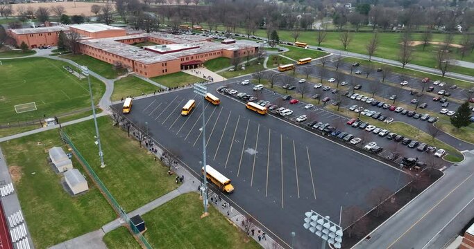 Aerial of American school campus with yellow school bus arrival and departure. Students walk on sidewalk by athletic field. Aerial view.
