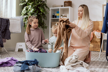 A woman and two daughters with blond and bronze hair fold clean laundry taken out of the dryer, the...