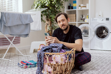 Portrait of man sitting in laundry room on floor by wicker basket of clothes with a phone in hand. The boy is sorting shirts checking the internet for pictograms, washing machine instructions.