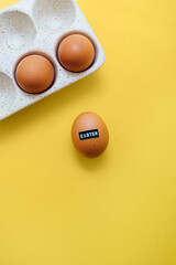 Brown raw chicken egg with embossed word Easter and eggs in a ceramic storage tray on a yellow background with copy space. Creative holiday decor and spring greeting card. Flat lay.