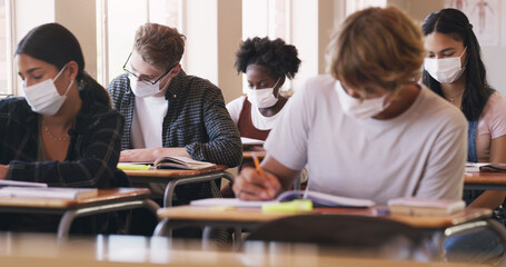Nothing gets in their way of getting that grade. Shot of masked teenagers writing an exam in a...
